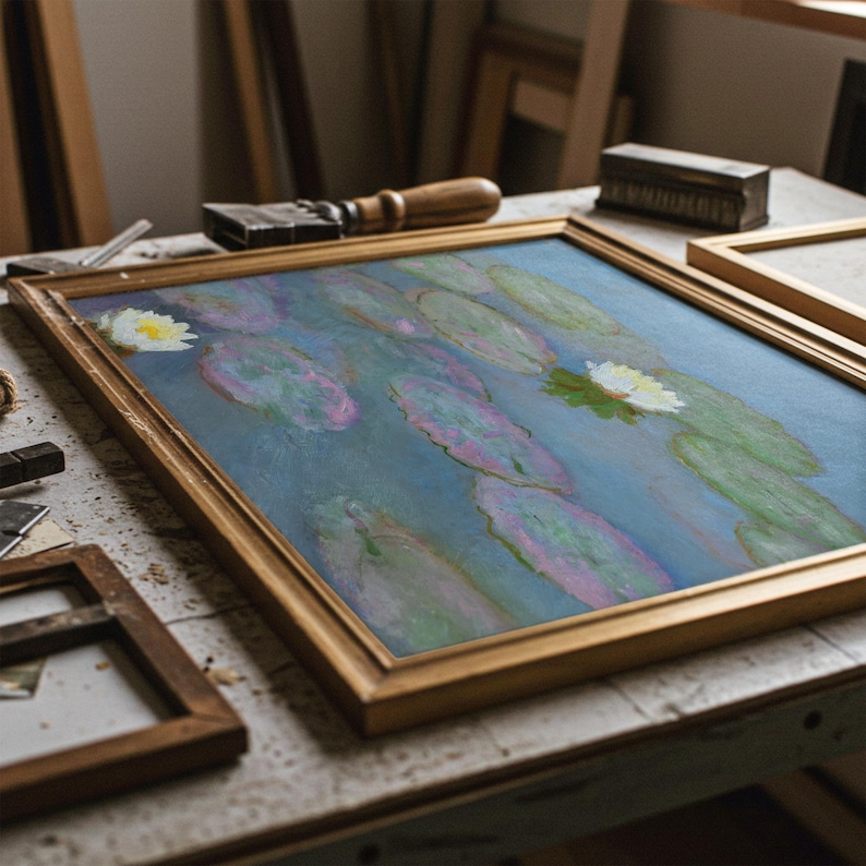 May include: A framed painting of water lilies in a pond, with a gold-colored wooden frame. The artwork features a blue background with green lily pads and white flowers. Tools and other frames are visible on the work surface.