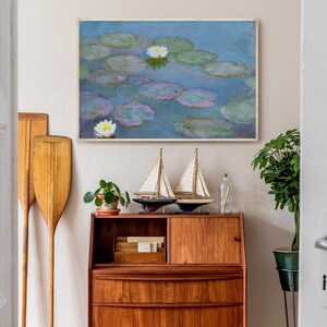May include: A framed painting of water lilies in a pond hangs above a wooden cabinet. Two decorative sailboat models and a small potted plant sit on top of the cabinet. Two wooden oars lean against the wall.