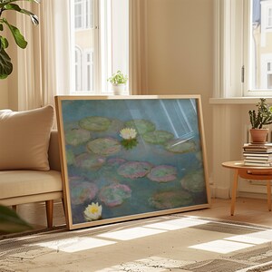 May include: A framed print of a water lily pond scene. The artwork features lily pads in shades of green and pink, with white water lilies. The frame is a light wood color, and the print is displayed indoors.