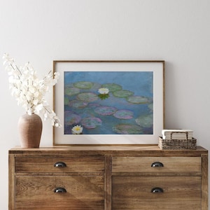 May include: A framed print of a water lily pond, featuring lily pads and white flowers, in a wooden frame. The artwork is displayed on a wooden dresser, with a vase of white flowers and a small woven basket.