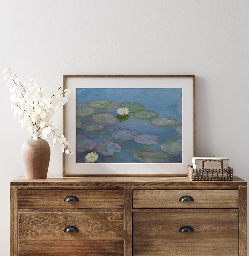 May include: A framed print of a water lily pond, featuring lily pads and white flowers, in a wooden frame. The artwork is displayed on a wooden dresser, with a vase of white flowers and a small woven basket.