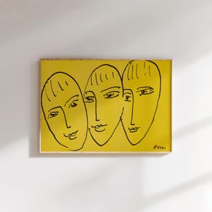 Peut inclure: Une impression d'art encadrée représentant trois visages stylisés à l'encre noire sur fond jaune. L'œuvre a un style minimaliste avec des lignes et des formes simples. Le cadre est de couleur bois clair.