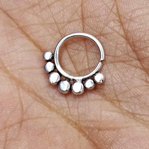 Könnte beinhalten: Ein silberner Septumring mit einer Reihe kleiner, runder, polierter Perlen. Der Ring ist kreisförmig mit einer Scharnieröffnung. Die Perlen sind gleichmäßig auf der unteren Hälfte angeordnet und bilden einen dekorativen Akzent.