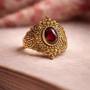 Könnte beinhalten: Verzierter goldfarbener Ring mit einem großen, ovalen, tiefroten Edelstein in der Mitte. Der Ring hat aufwendige Schnörkel und Perlenverzierungen um den Edelstein und das Band. Ein Schmuckstück im Vintage-Stil.