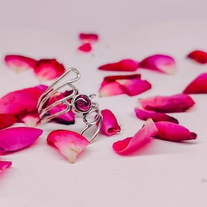 Könnte beinhalten: Ein silberfarbener Ring mit einem offenen, mehrschichtigen Drahtbanddesign, das sich um einen zentralen, kreisförmigen, dunkelmagentafarbenen Edelstein windet. Der Ring ist von verstreuten, leuchtend rosa und roten Rosenblättern umgeben.