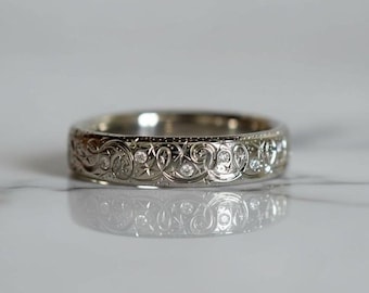 May include: A silver ring with an intricate engraved pattern of swirling designs and small, sparkling gemstones. The ring is displayed on a reflective surface, showcasing its detailed craftsmanship and elegant style.
