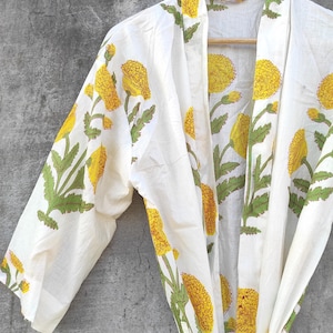Op de afbeelding: Witte kimono met een geel bloemenpatroon en groene bladeren, hangend aan een houten hanger. De kimono heeft een striksluiting en is gemaakt van een lichte stof. Het bloemmotief herhaalt zich over het hele oppervlak.