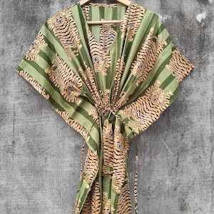 May include: A green and tan patterned kaftan dress with a V-neck and tie closure. The dress features a repeating tiger print and vertical stripes. The garment is hanging on a wooden hanger against a gray wall.