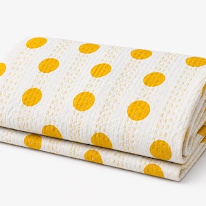 May include: Folded white blanket with large yellow polka dots. The blanket is made of a textured fabric and is folded neatly, showcasing the pattern. The blanket is a home textile.