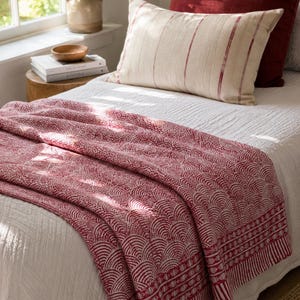 Peut inclure: Un lit avec une couette blanche, orné d'une couverture à motifs rouges et blancs. Deux oreillers sont visibles : un à rayures rouges et un autre rouge uni. Une table de chevet en bois contient des livres et un bol décoratif.