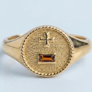 Pode incluir: Um anel de sinete dourado com uma face circular e uma borda em estilo de corda. O centro exibe uma cruz em relevo acima do texto "QUI HIER" e uma pedra retangular cor de âmbar acima do texto "QUI DEMAIN".
