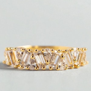 May include: A gold ring with a series of clear, geometric-cut gemstones. The gemstones alternate between rectangular and pear shapes, set in a gold band. The ring is displayed on a light gray surface.