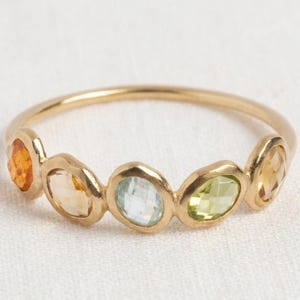 Peut inclure: Une bague en or ornée de cinq pierres précieuses ovales à facettes disposées en rangée. Les pierres présentent un dégradé de couleurs incluant orange, jaune pâle, bleu clair, vert citron et jaune doré, montées sur un anneau en or fin poli.