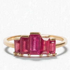Puede incluir: Un anillo de oro con cinco gemas rosas rectangulares y vibrantes. Las gemas están dispuestas en una fila, con la piedra central ligeramente más grande. El anillo tiene un diseño simple y elegante.