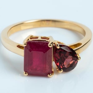 May include: A gold ring featuring two prominent red gemstones. One stone is cut in a rectangular emerald shape, while the adjacent stone is cut in a teardrop pear shape. The band is smooth, polished yellow gold, creating a simple and elegant setting.