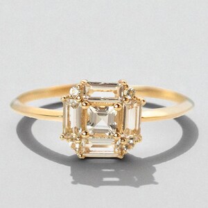 May include: A gold ring featuring a central square-cut gemstone surrounded by smaller rectangular and round stones. The ring has a delicate band and a cluster design around the main stone, creating a vintage or art deco style. The ring is set against a light gray background.
