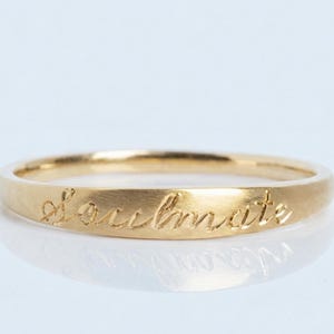 May include: A gold-colored ring with the word "soulmate" engraved on the band. The ring has a smooth, matte finish and is set against a white background. The ring is a simple band design.
