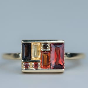 May include: A gold ring featuring a rectangular face set with a cluster of multi-colored gemstones. The arrangement includes a dark brown baguette, a light yellow baguette, a small dark round stone, two small red round stones, and two orange-red baguettes.