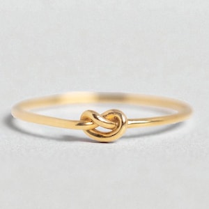May include: A delicate gold-colored ring featuring a knot design. The ring has a thin band and a small, detailed knot as the focal point. The ring is shown on a white background, highlighting its simple elegance.