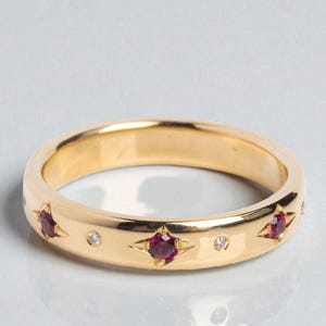 May include: A gold band ring with star-shaped settings, each holding a red gemstone. Small, round, clear gemstones are interspersed between the stars. The ring is displayed on a reflective surface.