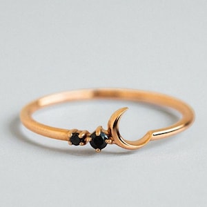 Puede incluir: Un anillo de oro rosa con un diseño de media luna y dos pequeñas piedras negras. El anillo tiene una banda delicada y un diseño minimalista, adecuado para el uso diario.