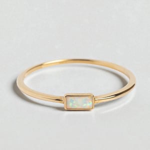 May include: A delicate gold ring featuring a rectangular opal stone. The ring has a thin band and a simple, elegant design. The opal stone has a white and iridescent appearance.