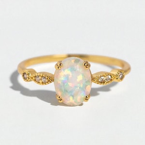 Può includere: Un anello d'oro con una pietra preziosa di opale ovale. L'opale presenta colori iridescenti. La fascia d'oro ha piccoli accenti decorativi su ogni lato dell'opale, ognuno con piccole gemme trasparenti.