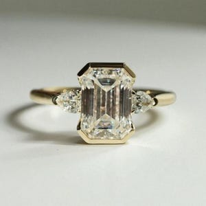 May include: A gold ring featuring a large, rectangular emerald-cut diamond as the centerpiece. Two smaller pear-shaped diamonds flank the main stone. The ring has a classic, elegant design.
