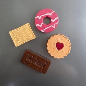 May include: Four biscuit-shaped magnets. Includes a Custard Cream, a pink donut with white zigzags, a heart-filled jammy dodger, and a chocolate Bourbon biscuit with the word "BOURBON" on it.