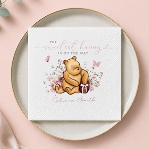 May include: A square white paper napkin featuring a printed illustration of a light brown bear holding a small honey pot. The text reads THE sweetest hunny IS ON THE WAY and Olivia Smith. Pink floral accents and butterflies surround the bear.