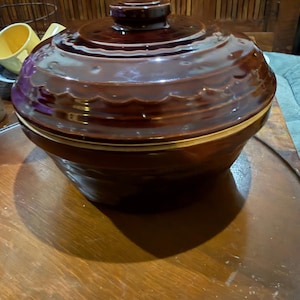 May include: A brown ceramic casserole dish with a lid. The dish has a glossy finish and a decorative wavy pattern around the rim. The lid has a small, round handle. The dish sits on a wooden surface.