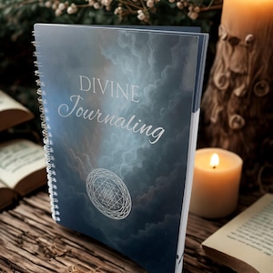 Puede incluir: Un diario con espiral con las palabras "DIVINE Journaling" en plata. La portada presenta un diseño azul nublado y un símbolo geométrico plateado. El diario está sobre una superficie de madera con velas encendidas y vegetación en el fondo.
