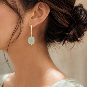 May include: A gold-toned earring featuring a single, smooth, rectangular light green stone bead suspended from a thin, slightly curved gold-toned wire hook. The earring hangs vertically from the earlobe, showcasing a minimalist design.