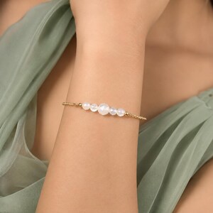May include: A delicate gold chain bracelet featuring a central row of five white, round, semi-translucent beads of varying sizes. The bracelet is secured with a thin, textured gold-toned chain that wraps around the wrist, creating a refined look.