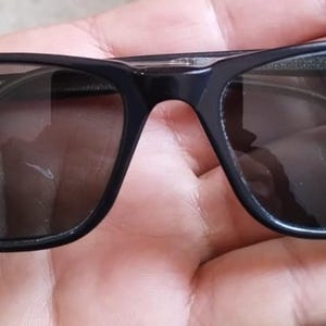 May include: Black rectangular sunglasses with dark tinted lenses. The sunglasses have a sleek, modern design with a subtle logo on the left arm. The frames are a glossy black, and the lenses are a dark gray color.