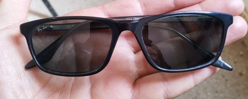 May include: Black rectangular sunglasses with dark tinted lenses. The sunglasses have a sleek, modern design with a subtle logo on the left arm. The frames are a glossy black, and the lenses are a dark gray color.
