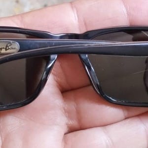 May include: Black sunglasses with dark lenses. The sunglasses have a rectangular frame and the Ray-Ban logo is visible on the top left of the frame. The arms of the sunglasses are folded.