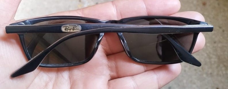 May include: Black sunglasses with dark lenses. The sunglasses have a rectangular frame and the Ray-Ban logo is visible on the top left of the frame. The arms of the sunglasses are folded.