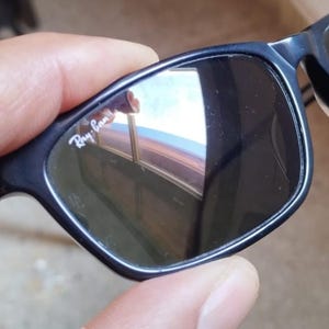 May include: Black sunglasses with dark lenses. The frames are a dark blue color. The Ray-Ban logo is visible on the upper left lens. The sunglasses are held by a hand.