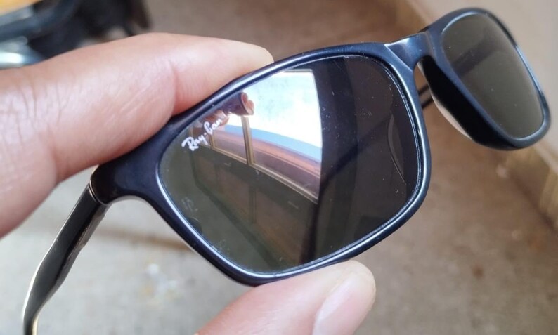 May include: Black sunglasses with dark lenses. The frames are a dark blue color. The Ray-Ban logo is visible on the upper left lens. The sunglasses are held by a hand.