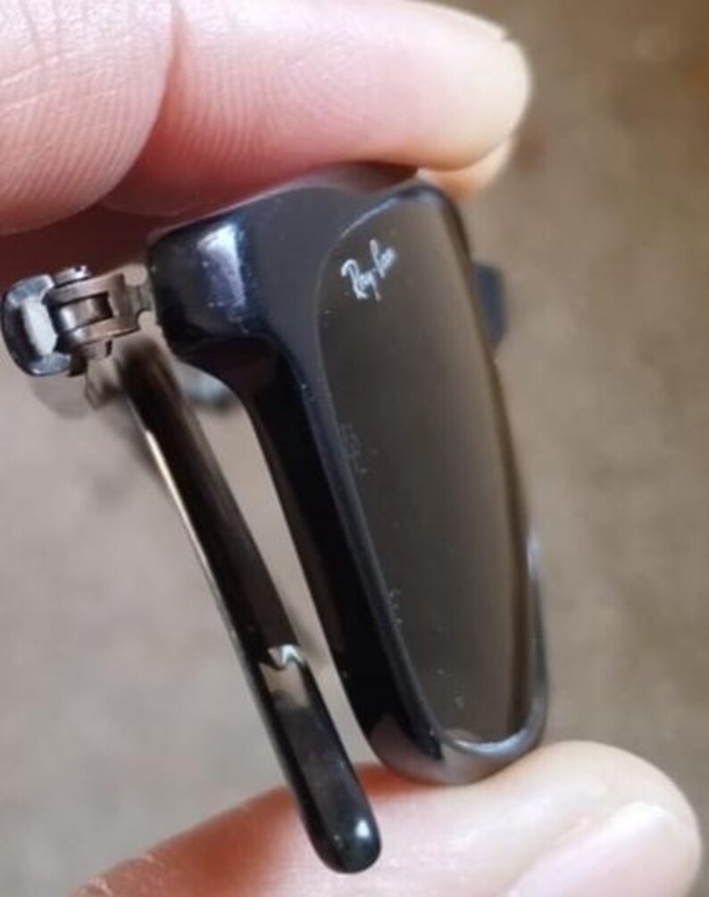 May include: Close-up of black sunglasses with the Ray-Ban logo in white. The sunglasses are partially folded, revealing the hinge and the sleek design of the frame and arms. The lenses are dark, and the overall look is modern.