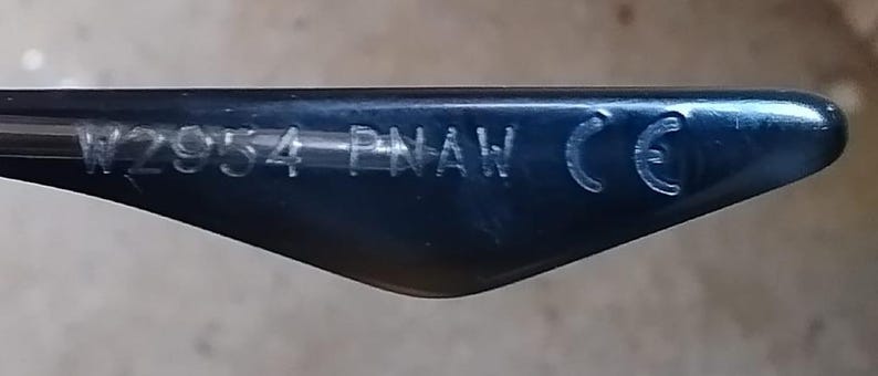 May include: Close-up of a dark blue eyeglass arm. The text "W2954 PNAW CE" is etched in white on the arm. The arm has a curved, triangular shape.