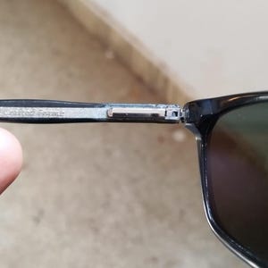 May include: Close-up of black sunglasses with a dark green lens. The frame is black with a silver hinge. The text "Real Crafted Frame" is etched on the arm of the sunglasses.