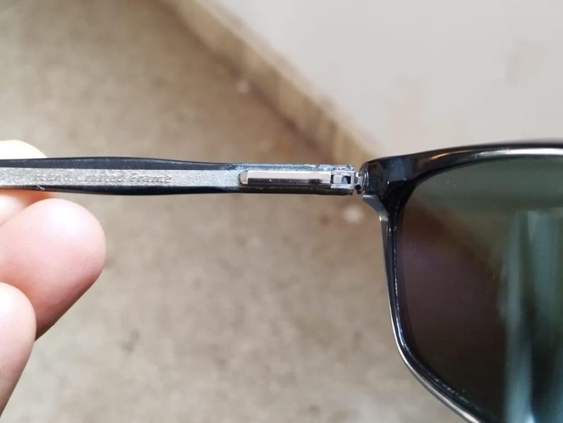 May include: Close-up of black sunglasses with a dark green lens. The frame is black with a silver hinge. The text "Real Crafted Frame" is etched on the arm of the sunglasses.