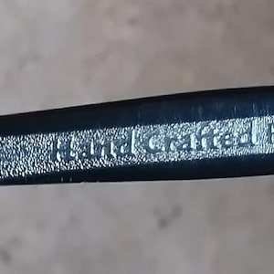 May include: Close-up of a black eyeglass frame with the words "Hand Crafted Frame" in silver lettering. The frame has a textured surface and a sleek, modern design.