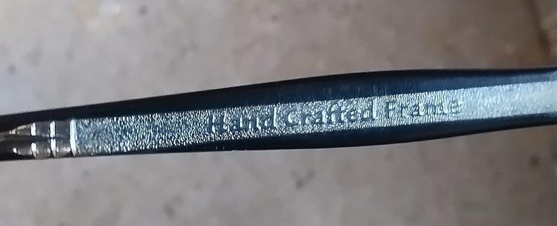 May include: Close-up of a black eyeglass frame with the words "Hand Crafted Frame" in silver lettering. The frame has a textured surface and a sleek, modern design.