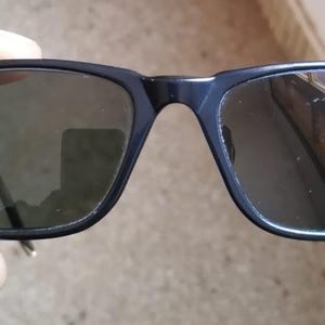 May include: Black rectangular sunglasses with dark lenses. The sunglasses have a matte finish and the Ray-Ban logo is visible on the left lens. The frames are held by a hand.