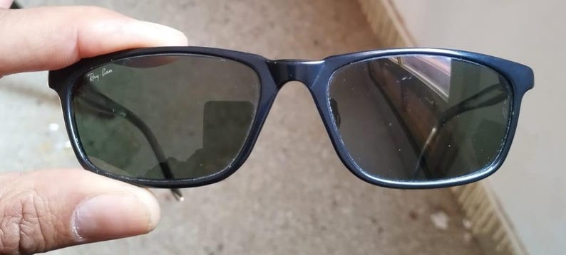 May include: Black rectangular sunglasses with dark lenses. The sunglasses have a matte finish and the Ray-Ban logo is visible on the left lens. The frames are held by a hand.