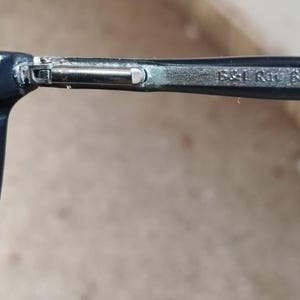 May include: Close-up of a pair of sunglasses with a dark blue frame and green lenses. The arm of the glasses has the text "B&L Ray 601" printed on it. The Ray-Ban logo is visible on the lens.