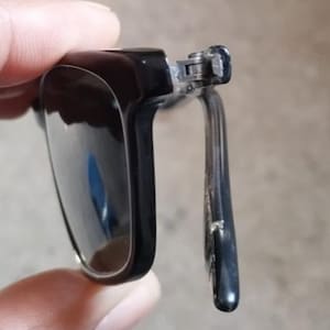 May include: Close-up of black sunglasses with a rectangular frame. The sunglasses have a dark lens and a silver hinge. The arm of the sunglasses is also black. The sunglasses are partially open.